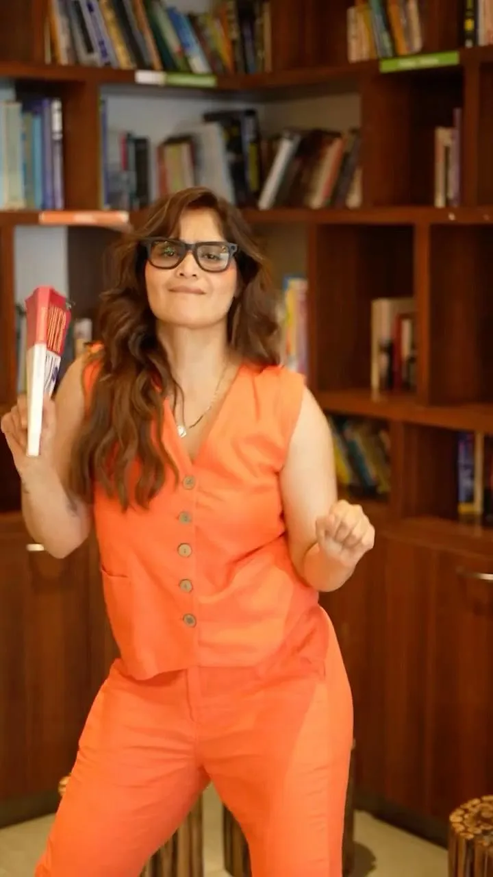 Arti Singh in an orange outfit standing by the bookshelves