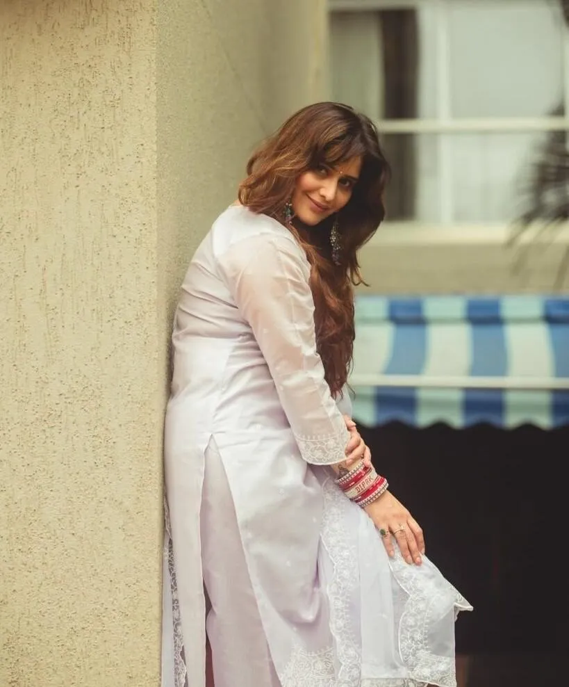 Arti Singh smiling softly in a white dress leaning on wall
