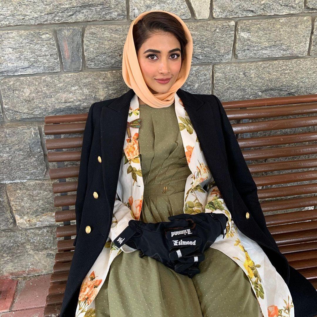 Pooja Hegde in Headscarf and Coat Sitting By Stone Wall