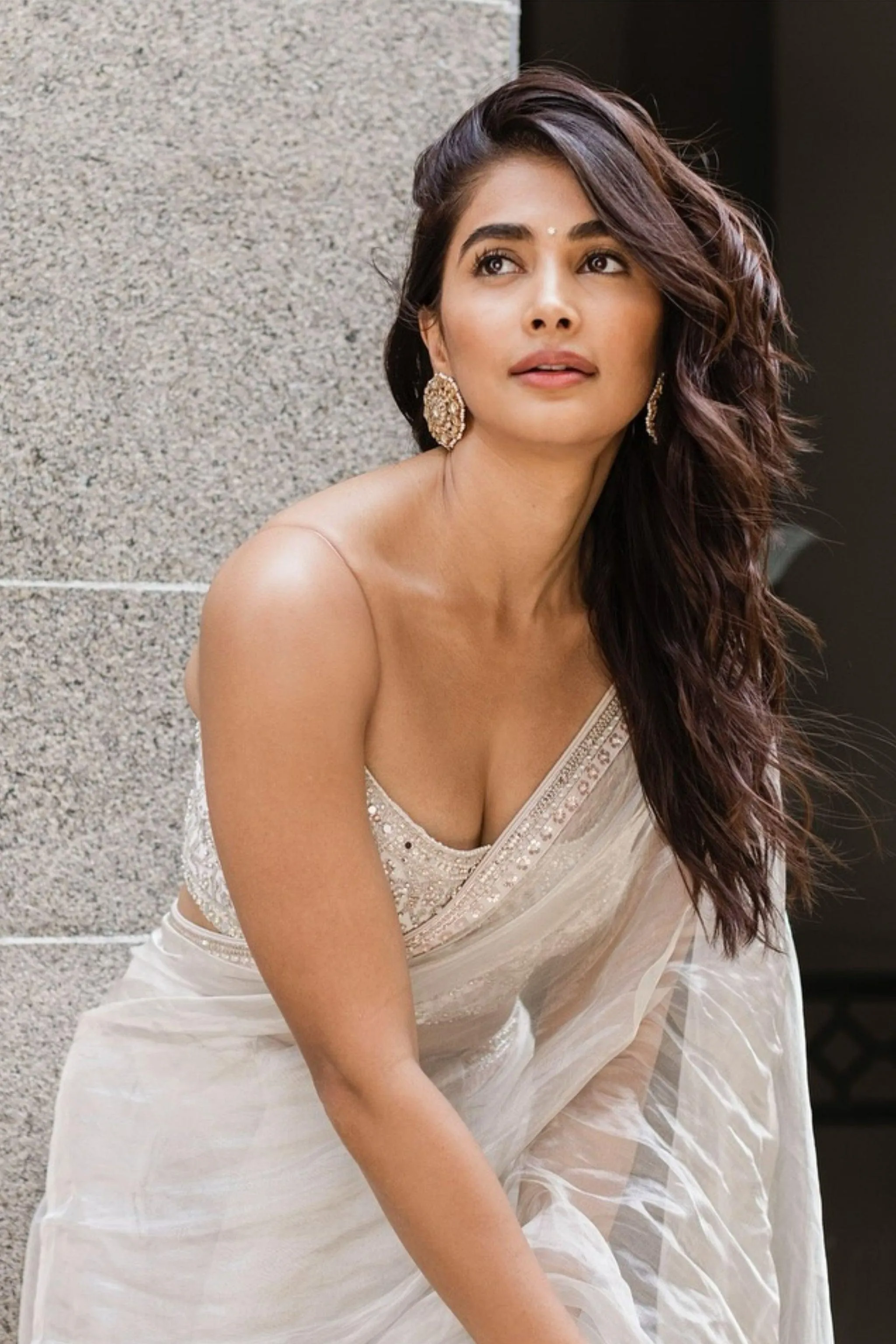 Pooja Hegde in Flowing White Dress Sitting Against Gray Wall