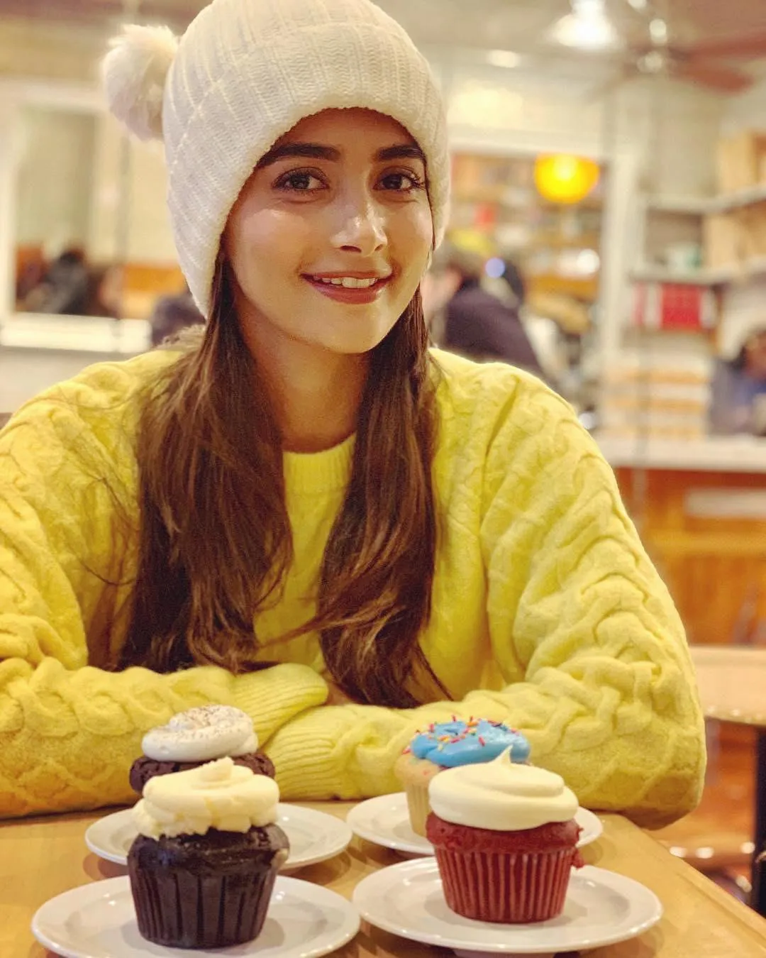 Pooja Hegde in Yellow Sweater Smiling with Cupcakes on Table