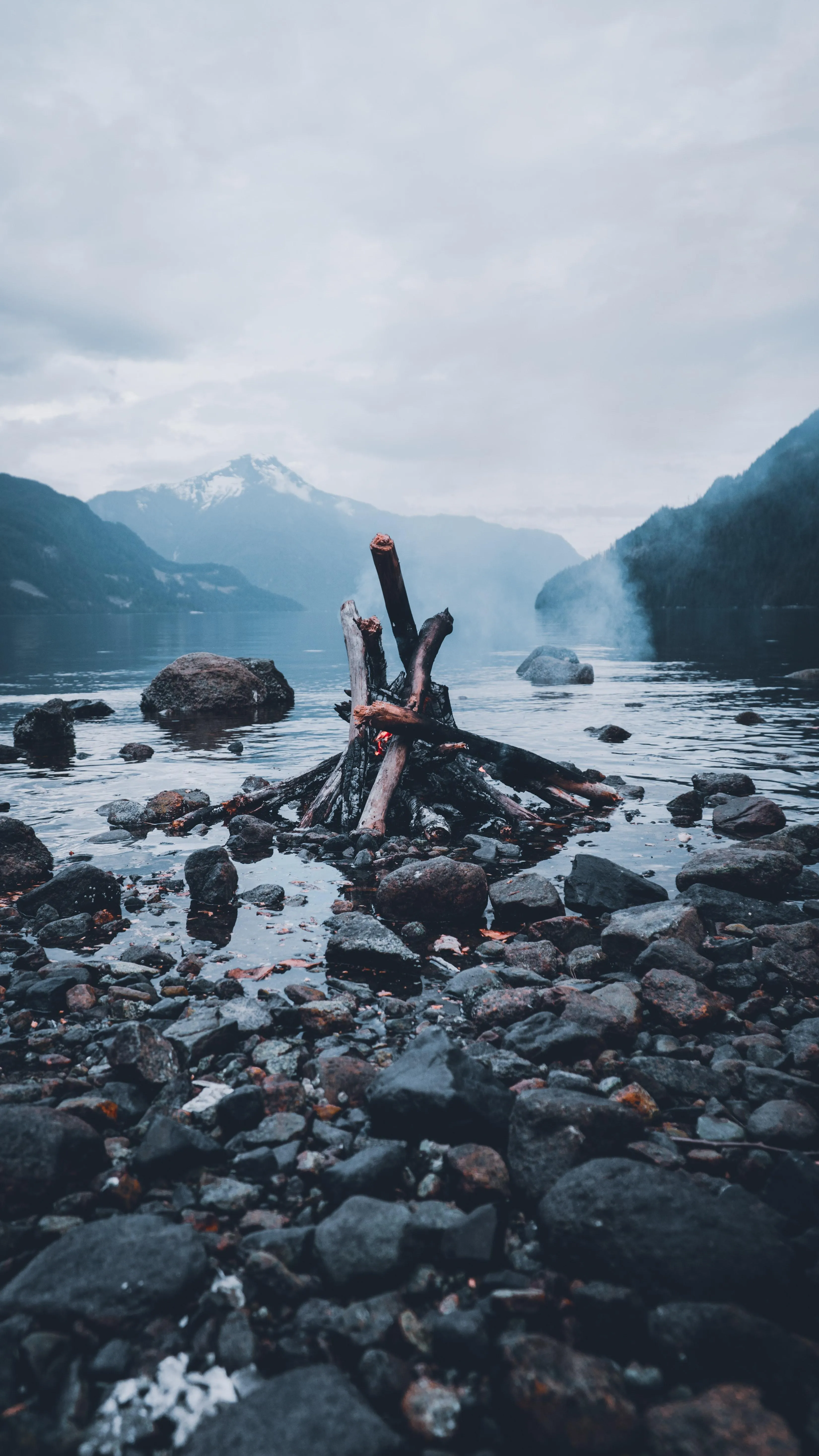 Campfire Beside Mountain Lake Under Overcast Sky Wallpaper