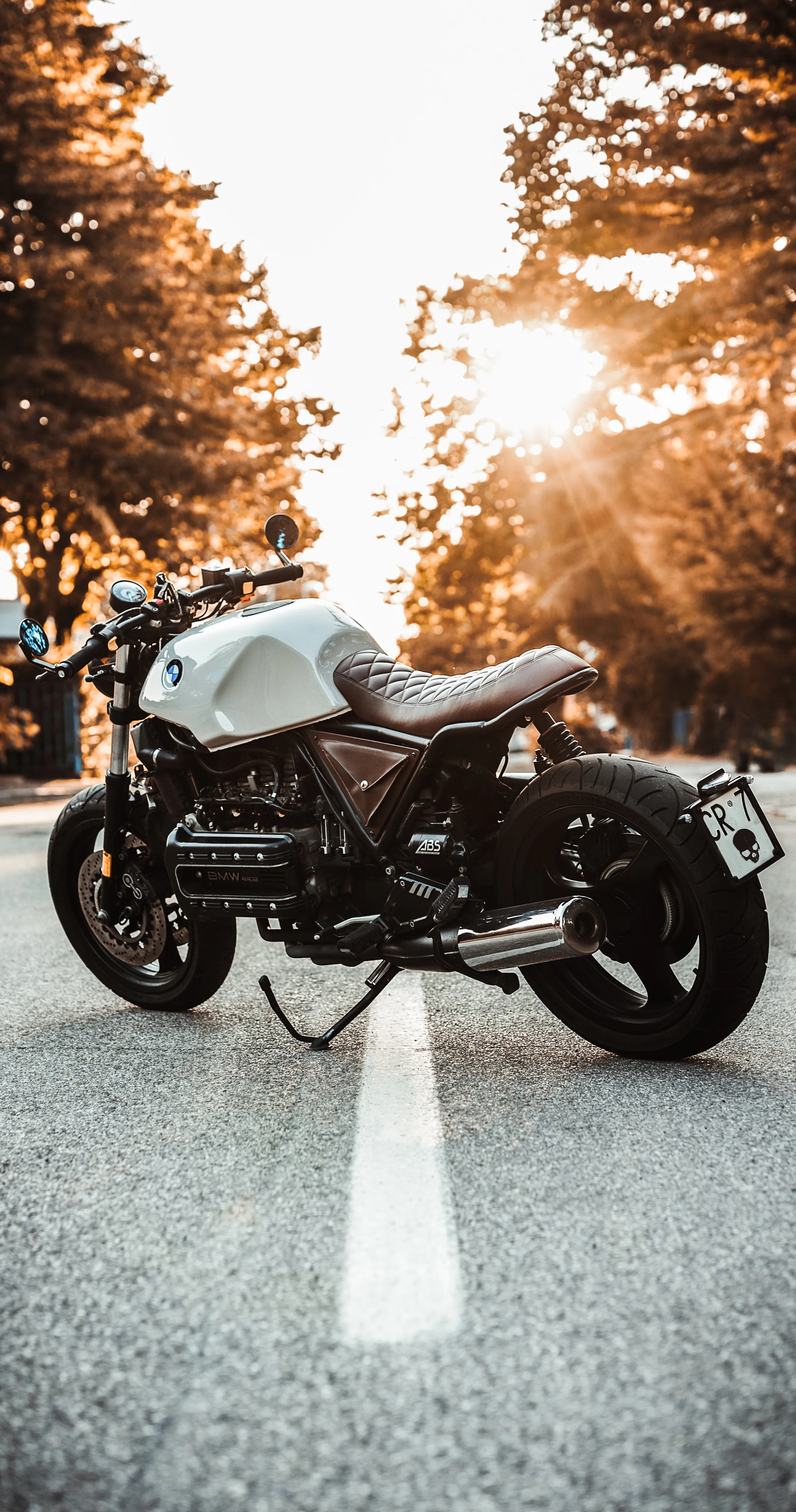 Classic Motorcycle Parked on Leafy Road at Sunset Wallpaper