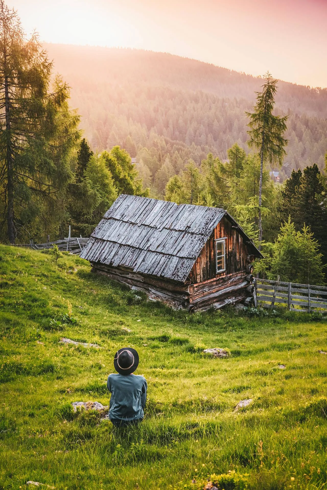Child sitting in a field near a wooden house and tall trees