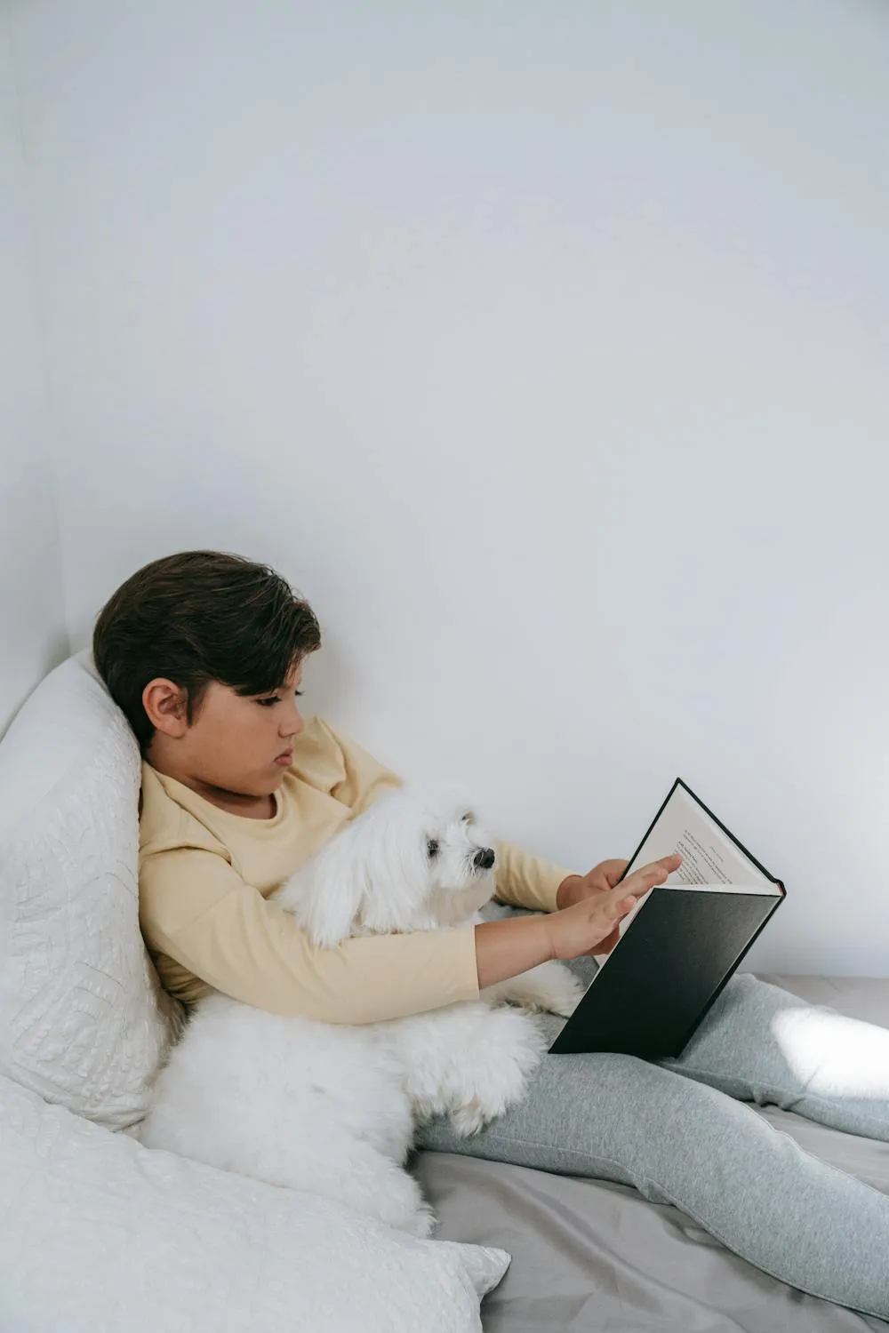 Boy Reading a Book in Bed While Hugging His White Dog