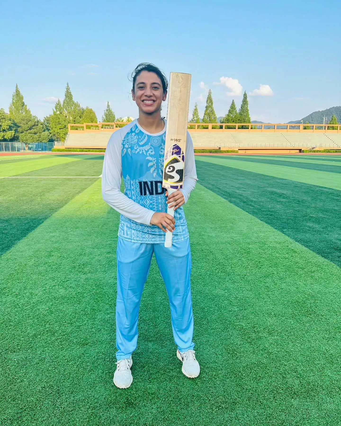 Smriti Mandhana posing in blue pants and white tee on a field