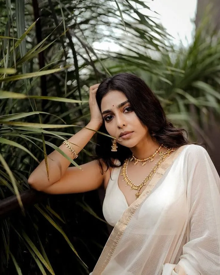 Aishwarya Lekshmi in a white saree sitting near the tall grass