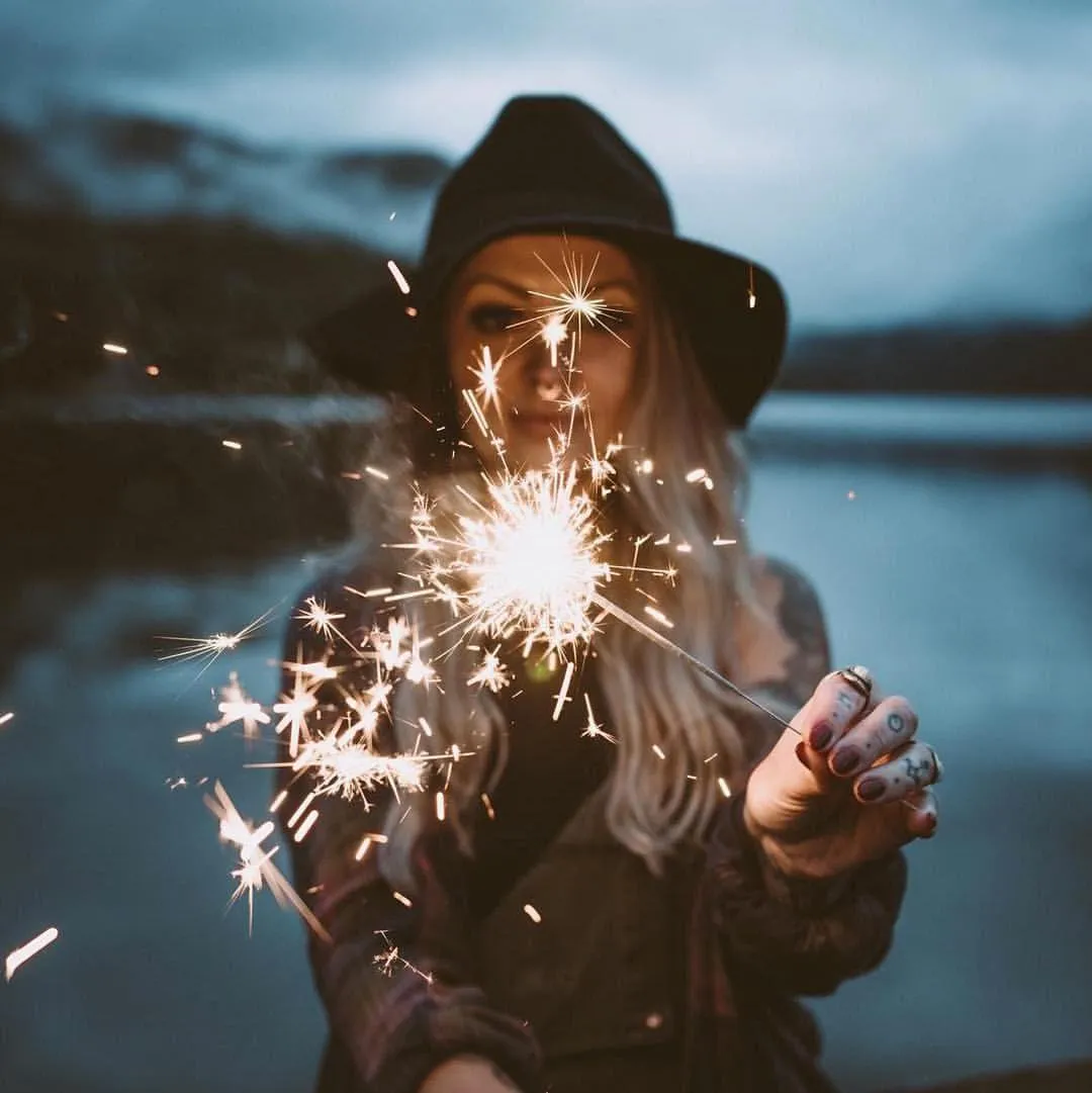 Girl Holding Sparklers Wearing a Black Hat Near Water Image