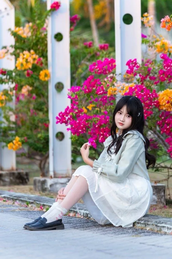 Girl in a White Dress Sitting Outside Near Bright Flowers