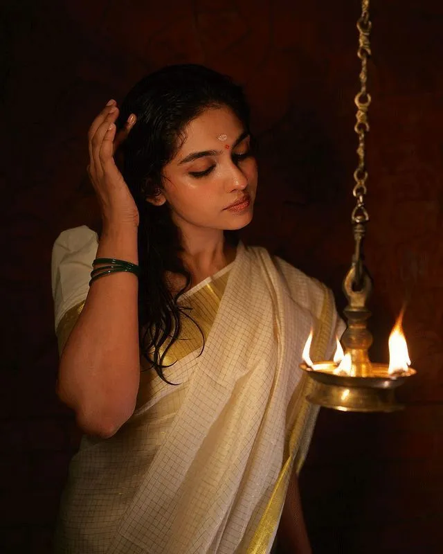 Mamitha Baiju in an Onam saree standing near the oil lamp
