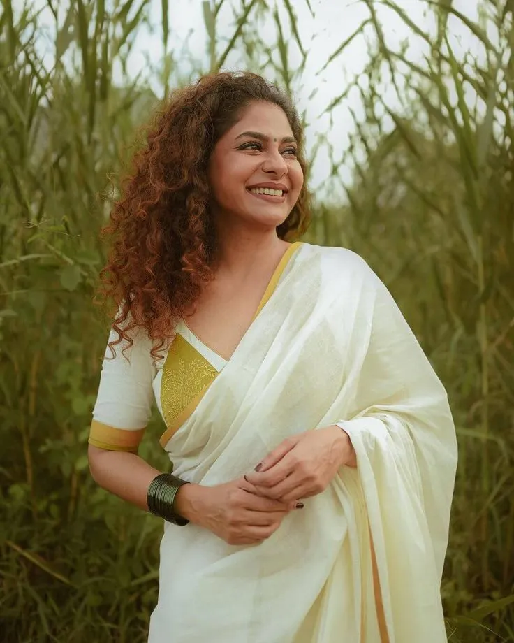 Poornima Indrajith in a Kerala saree near the tall grass