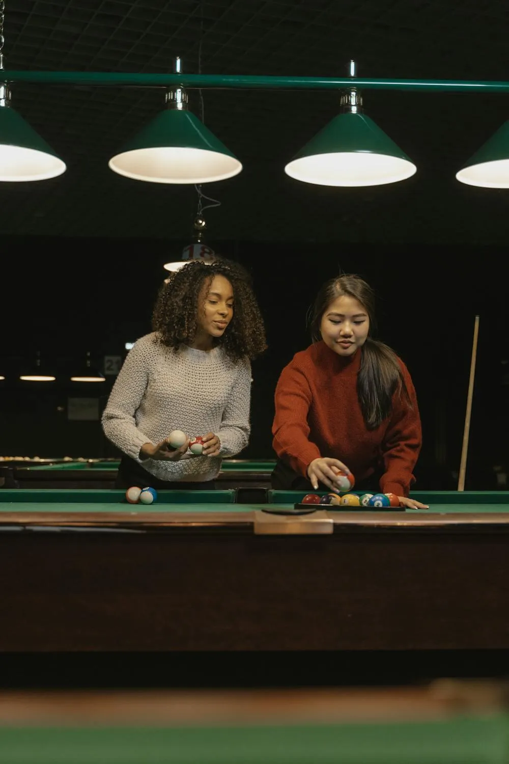 Two Women Playing Pool in Dimly Lit Room with Hanging Lights