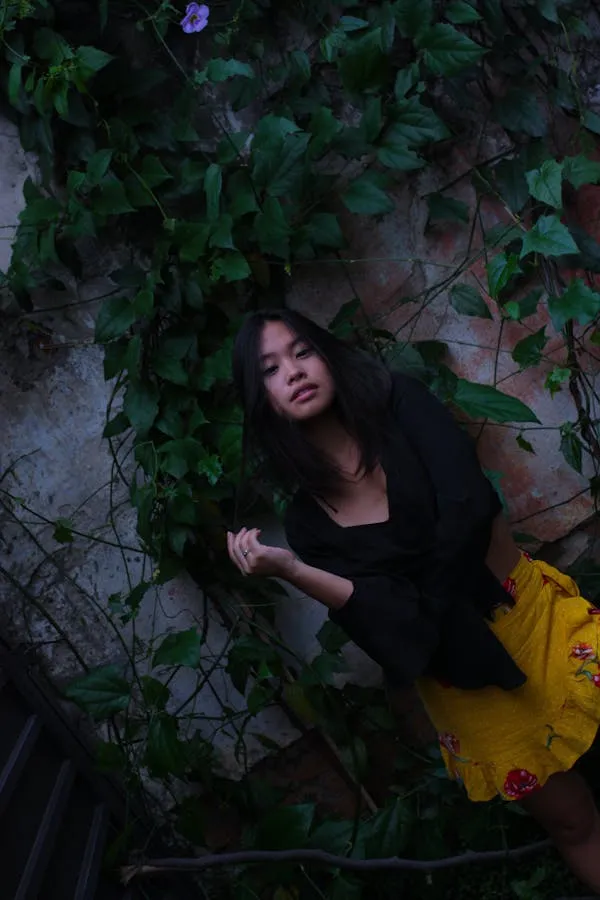 Woman in Black Top and Yellow Skirt Posing Next To Ivy Wall