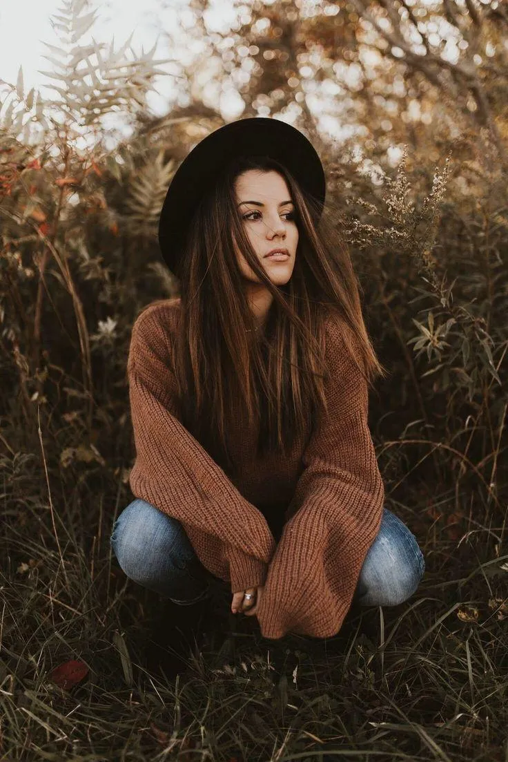 Woman Crouching in a Field Wearing a Brown Sweater and a Hat