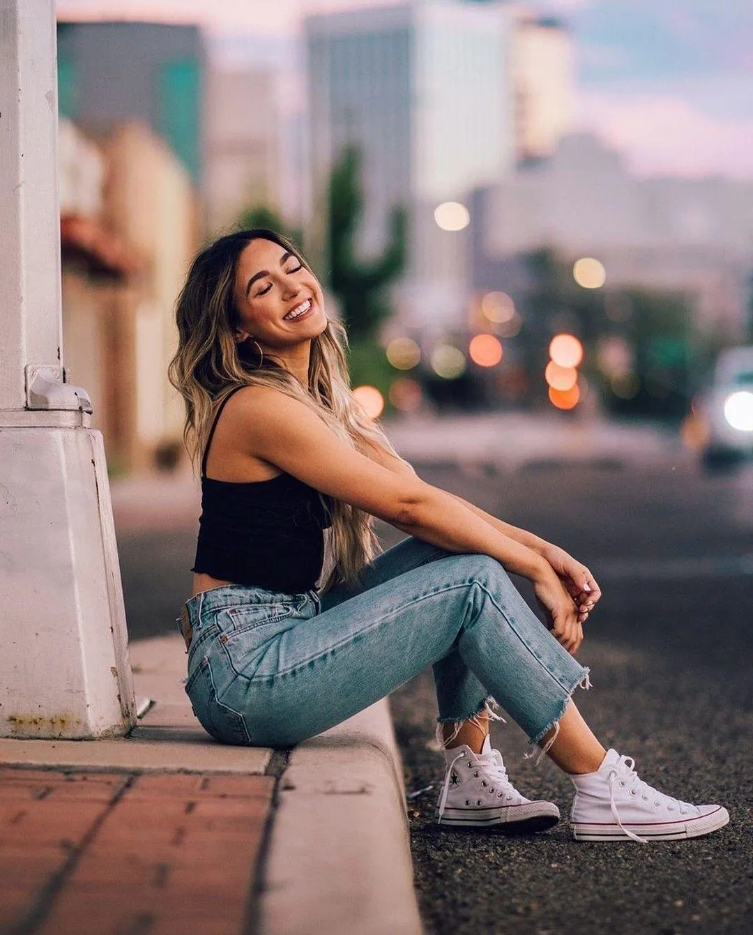 Woman Model Sitting on Sidewalk Smiling with City Background