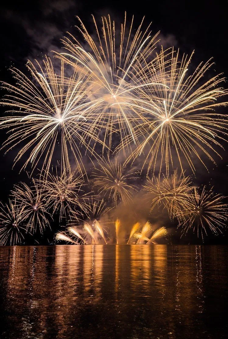 Bright golden fireworks reflect over a calm water bay