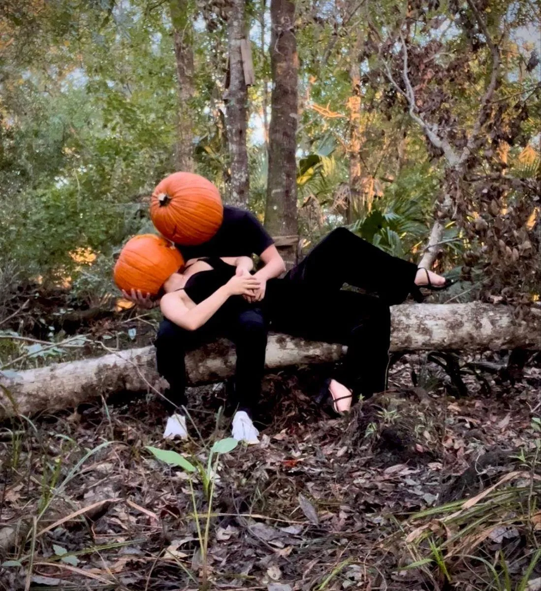 Two People in Pumpkin Masks Sitting on Log in Woods