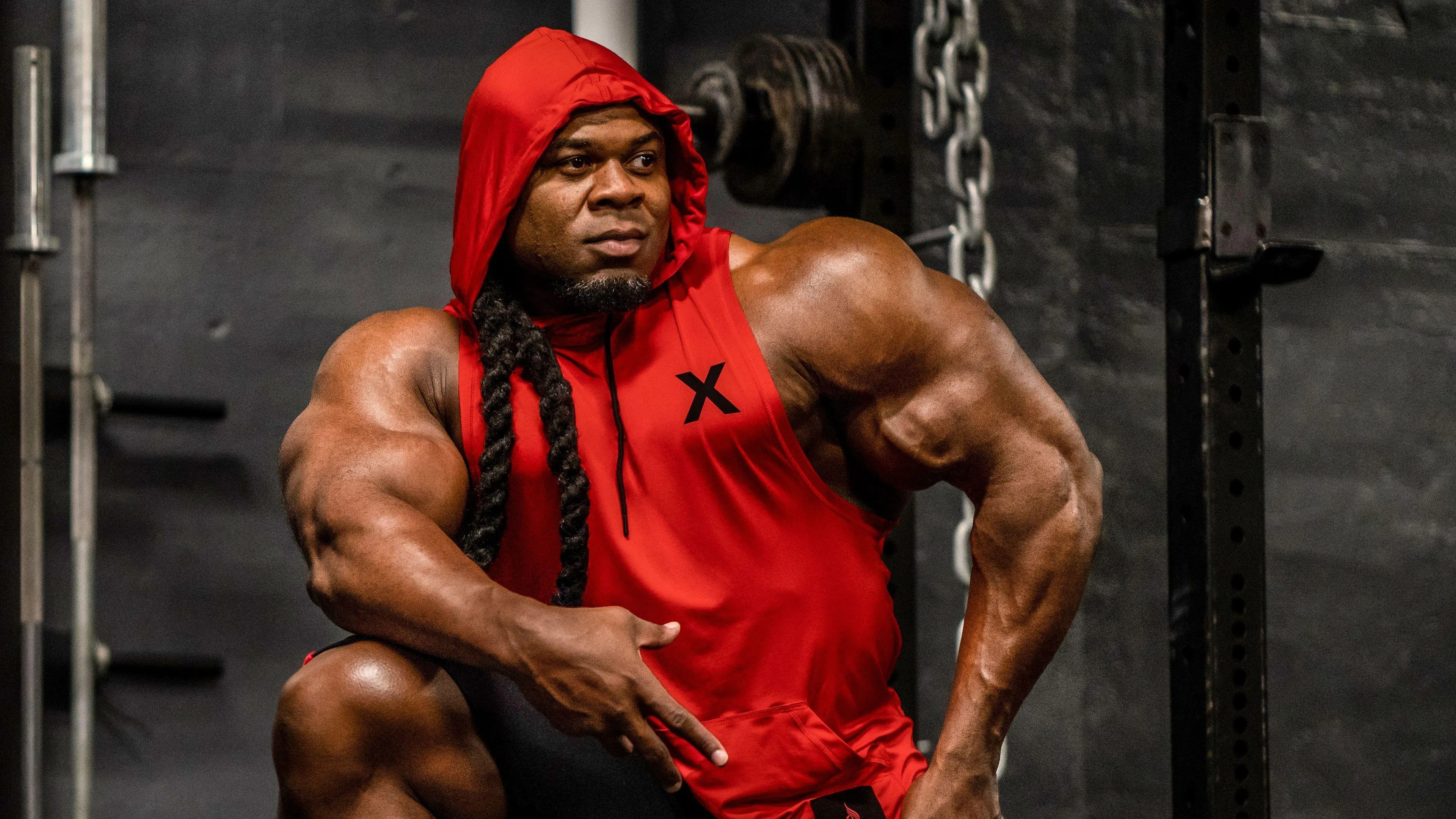 Kai Greene in a Red Hoodie Sitting Near the Gym Chains
