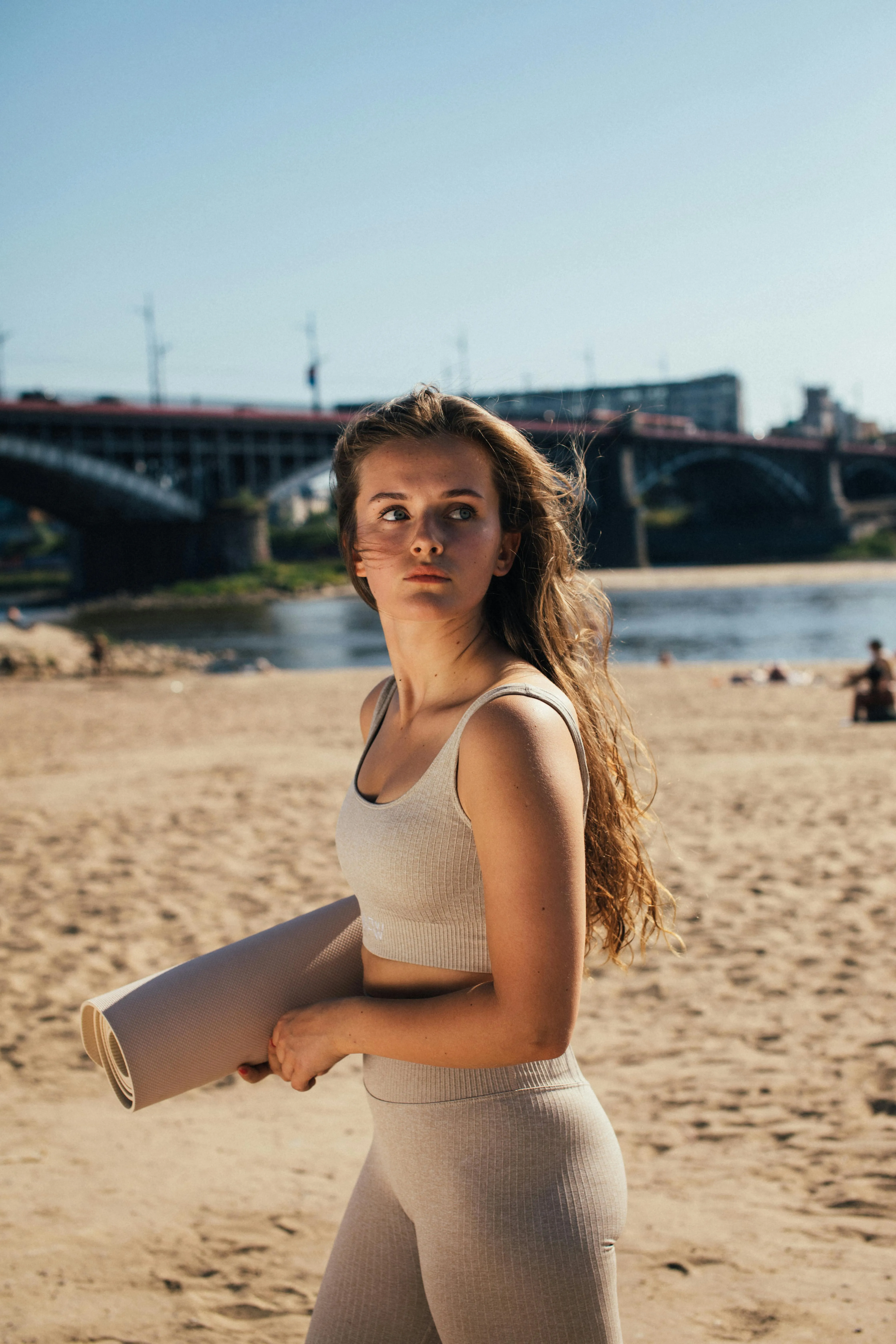 Woman Holding a Notebook Standing on the Beach Near a Bridge