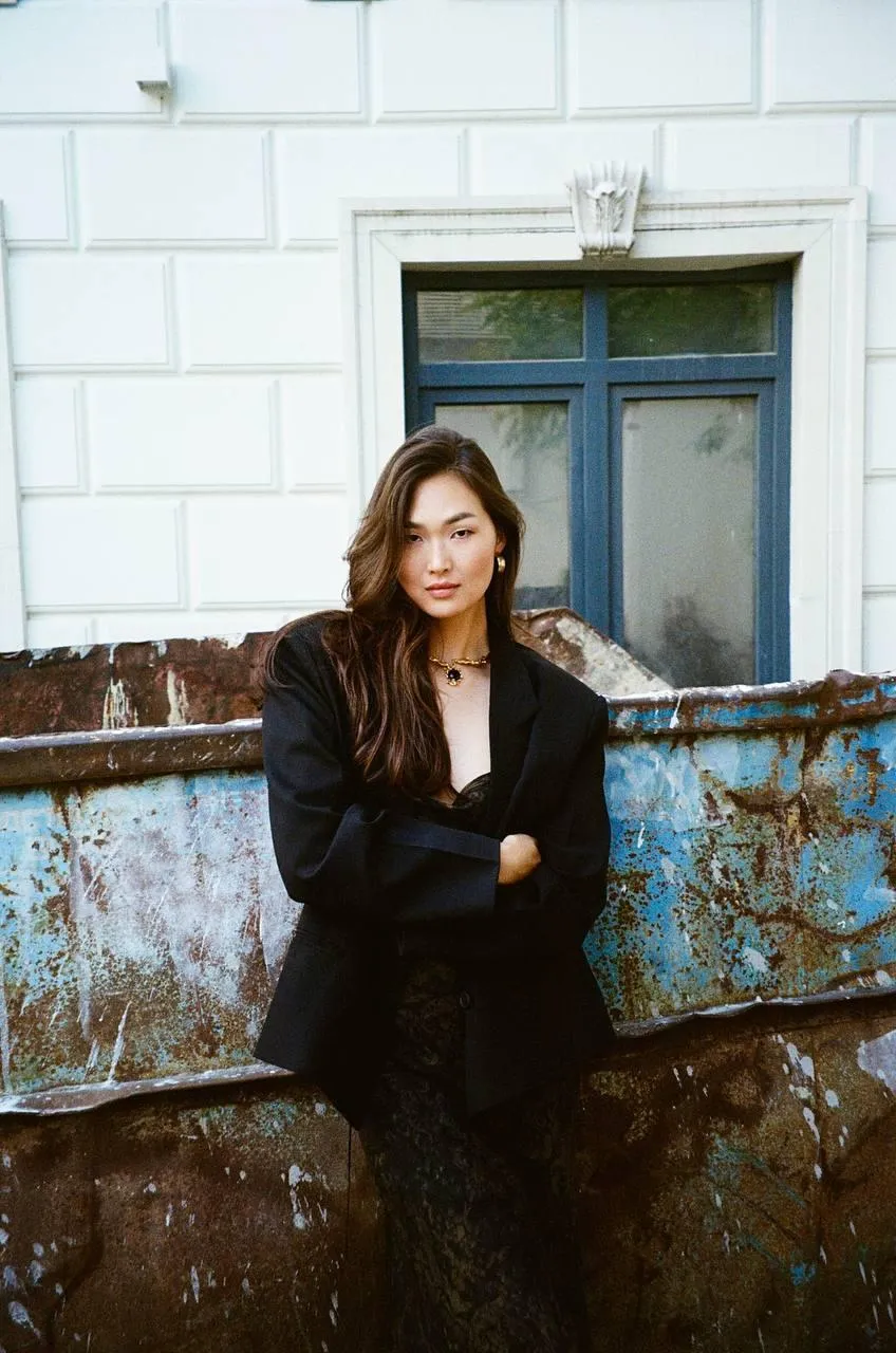 Woman in a Black Coat Sitting Against a Rustic White Wall