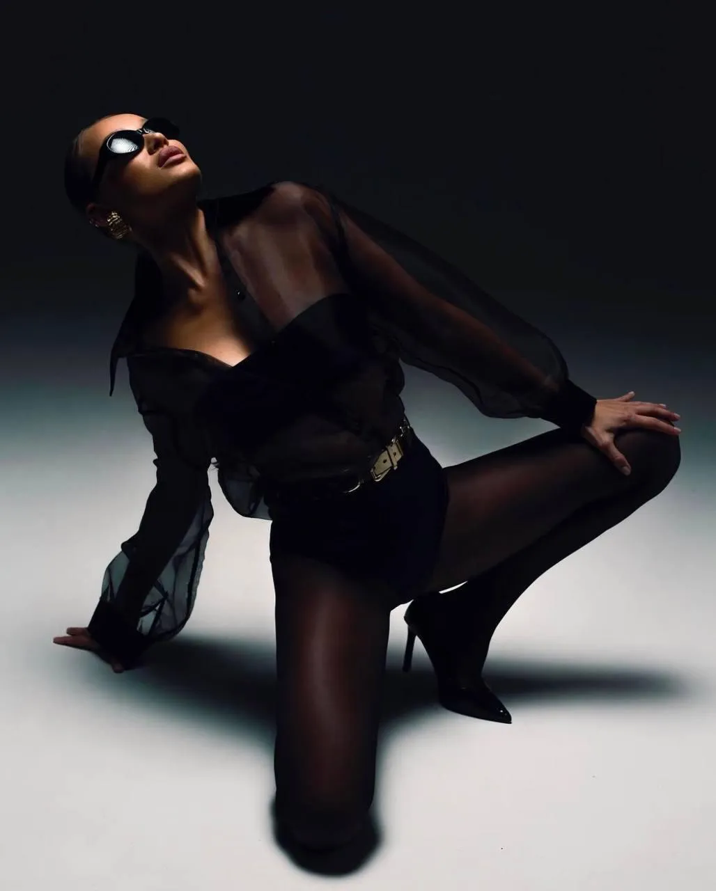 Woman in a black mesh dress and sunglasses kneeling on floor