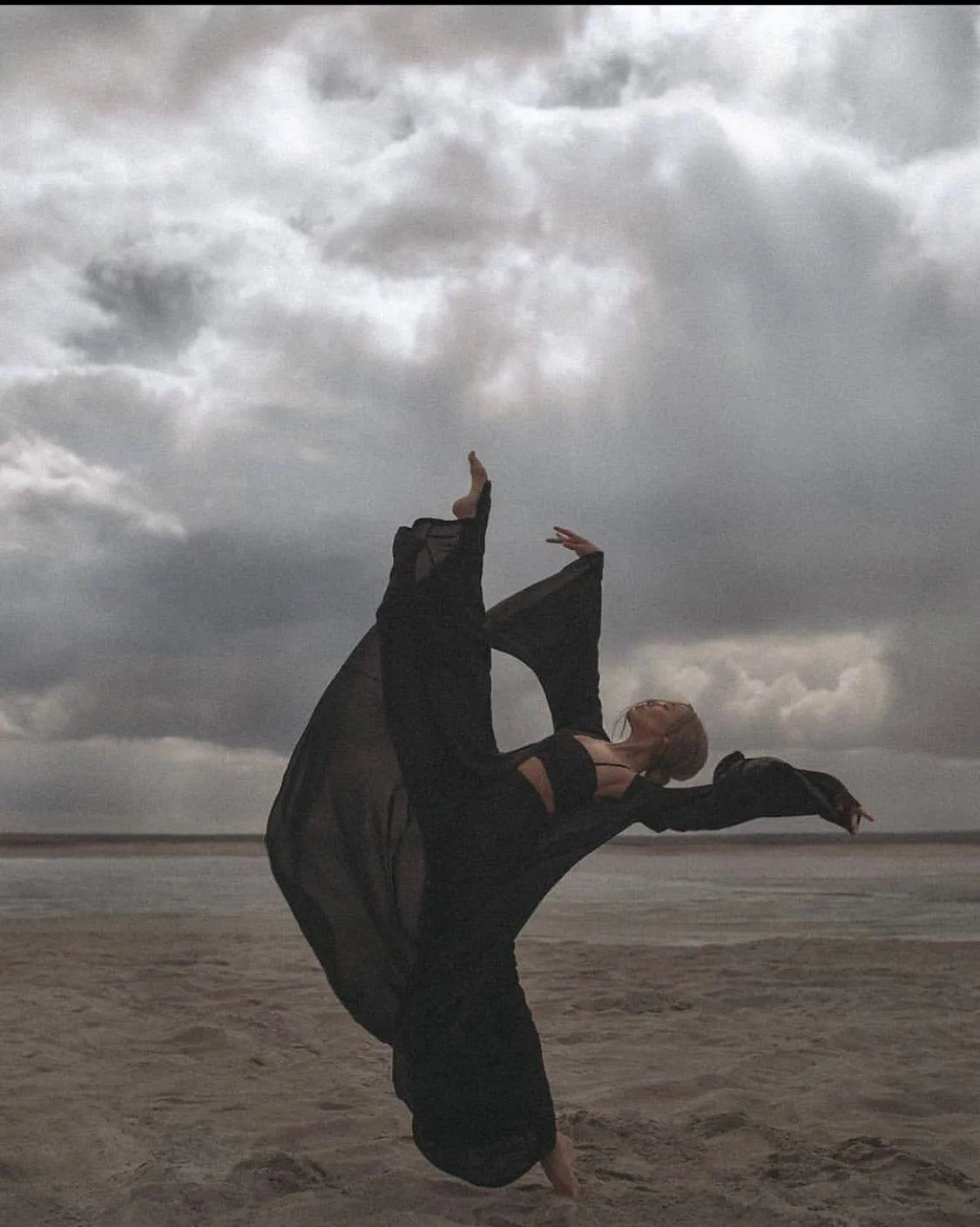 Woman in Black Flowing Dress Dancing on a Beach with Clouds