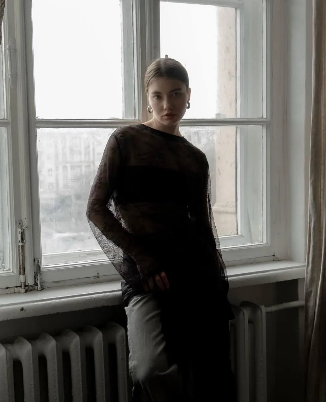 Woman Standing Near Window in Dark Outfit with Soft Shadows