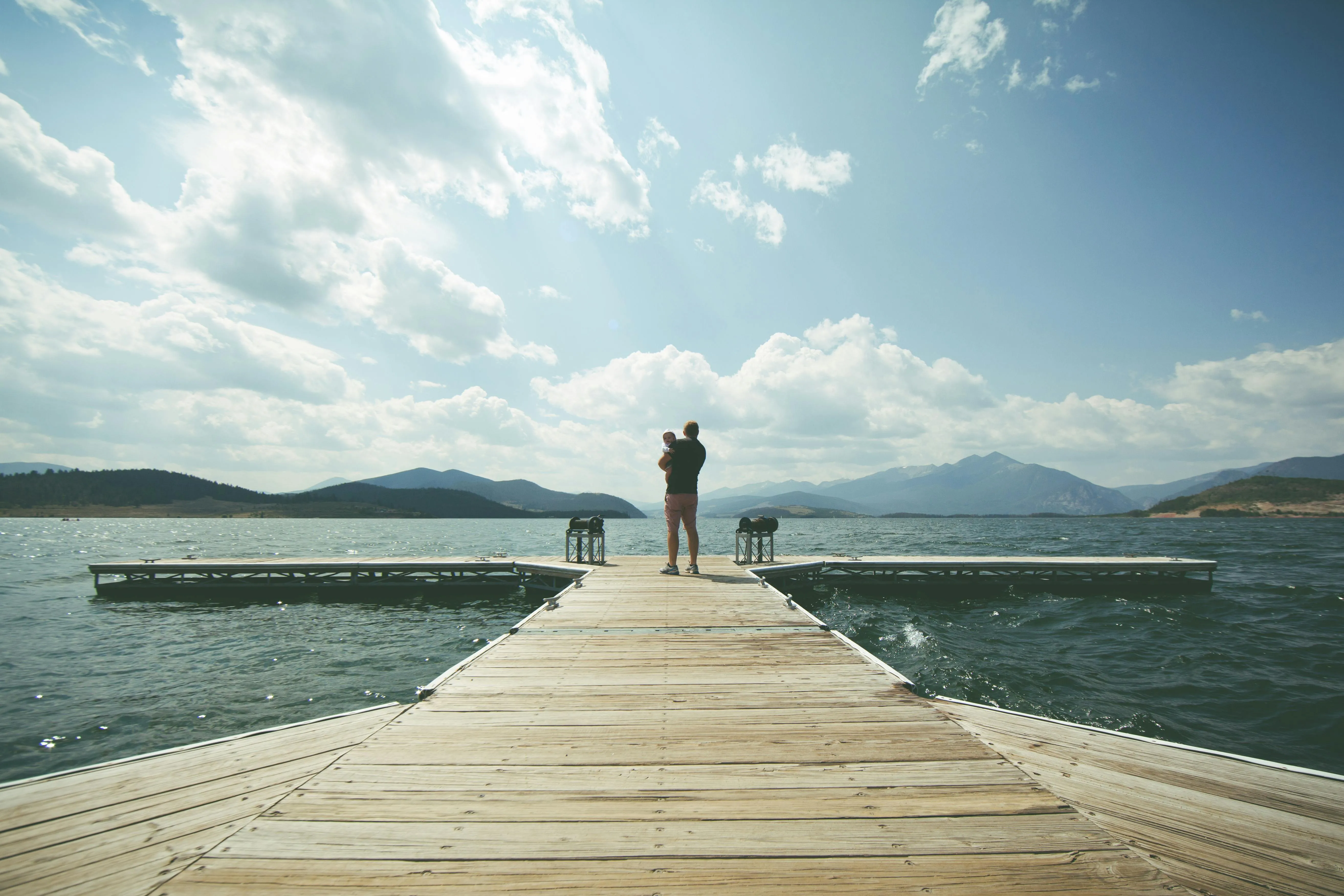 Man Standing on Pier Under Clear Sky and White Clouds