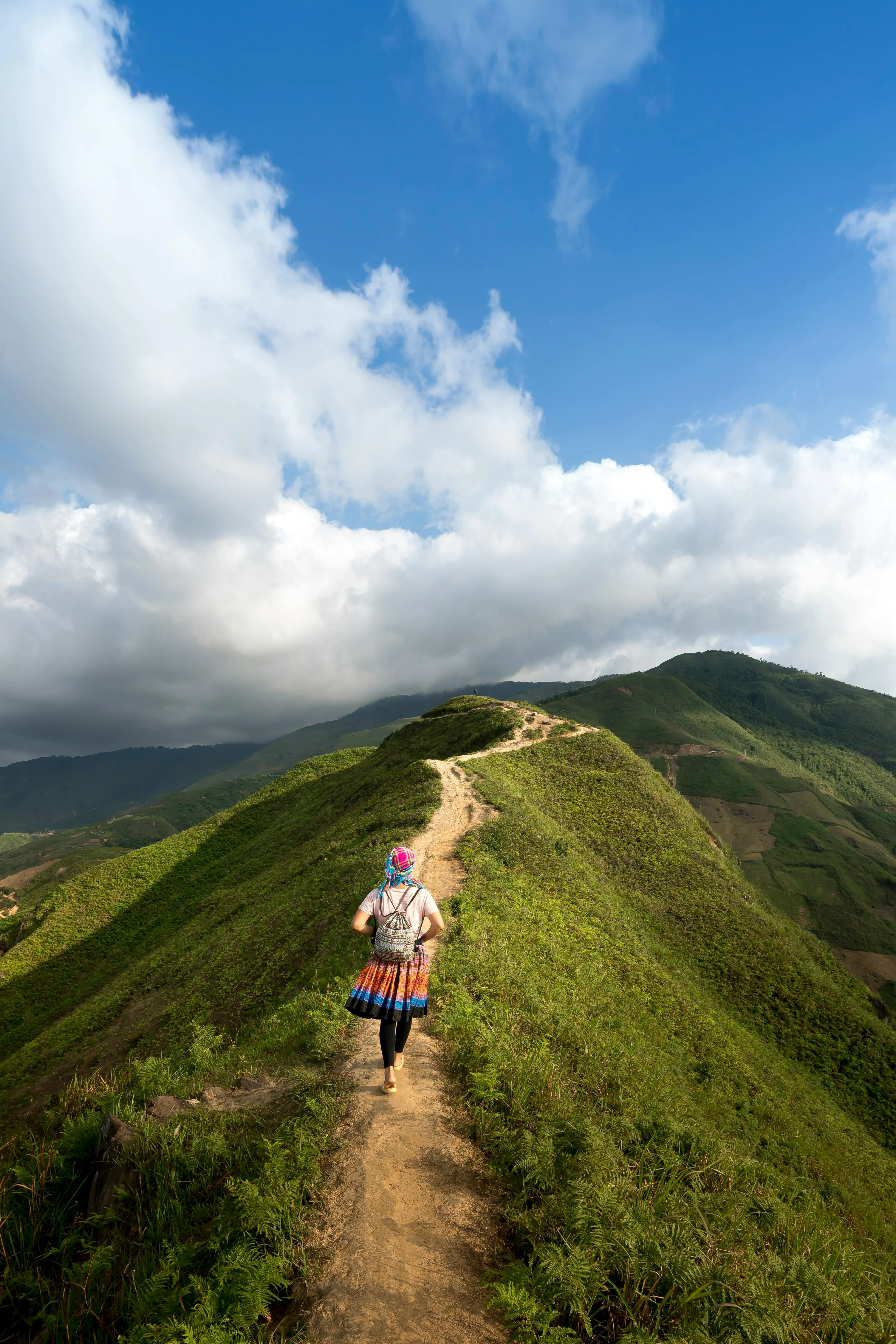 Person Walking on a Scenic Green Mountain Trail Under Clouds