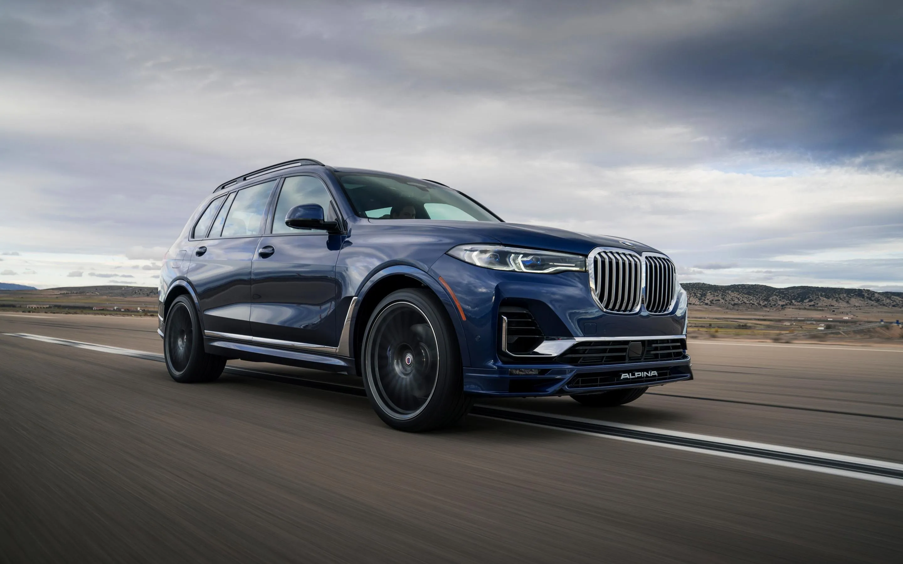Bmw Suv Driving on Dirt Road Under Cloudy Skies Wallpaper